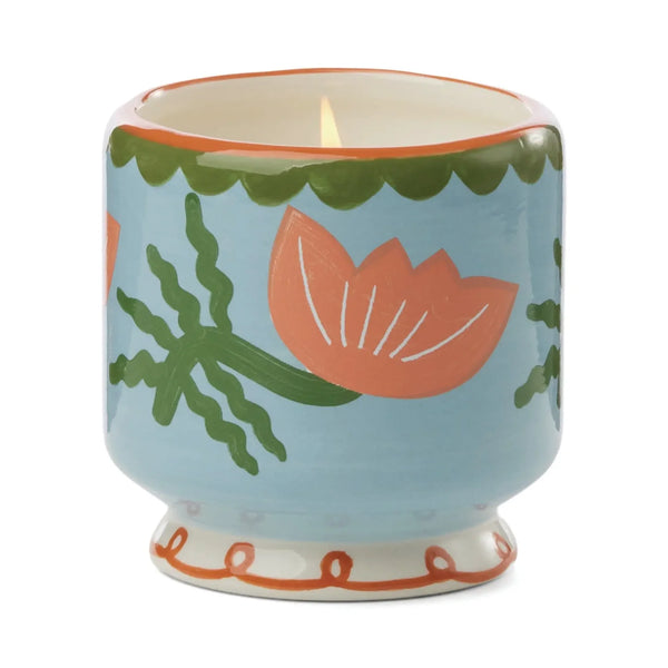 The A Dopo 8oz Ceramic Candle features a hand-painted design on a ceramic holder with a light blue background, pink flowers with green stems and leaves, a green scalloped rim, and a wavy red pattern at the base.