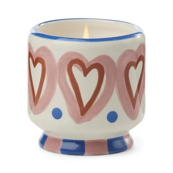 The A Dopo 8oz Ceramic Candle comes in a decorative ceramic holder with pink hearts, red outlines, blue dots, a blue and pink striped base, and is infused with a soothing Mediterranean fragrance.