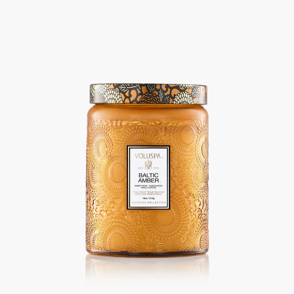 The Voluspa 18oz Glass Jar Candle features amber-colored embossed glass with floral patterns and a metallic lid, labeled 