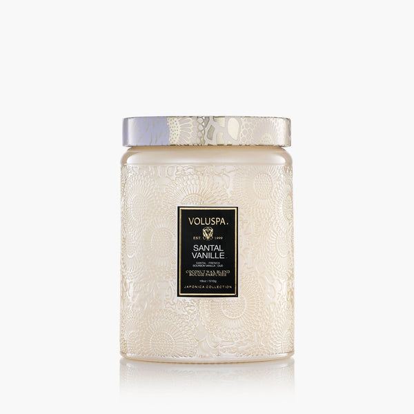 The Voluspa 18oz Glass Jar Candle features decorative white embossed glass with a matching lid. The label reads "Voluspa Santal Vanille, Japonica Collection" and the candle offers clean-burning performance with a minimalistic design.