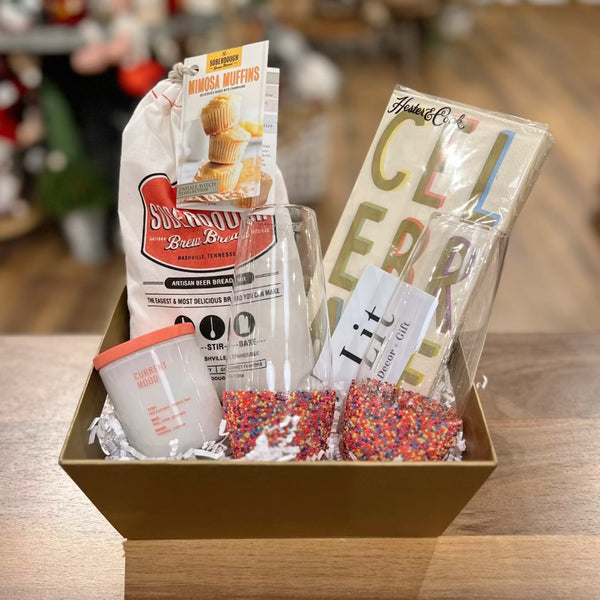 Create a Custom Gift Box from Lit Decor and Gift with a gold basket, mimosa muffin and bread mix, “Cheers” napkin set, “Clubber Mood” candle, matchbox, and two sprinkle tumblers—all arranged on a wooden surface.