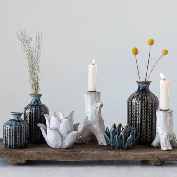 A wooden tray shows decorative vases, including a Blue Stoneware Vase with a striped pattern, dried plants, two white candles in tree trunk-shaped ceramic holders, and lotus-shaped tealight holders arranged against a plain background.