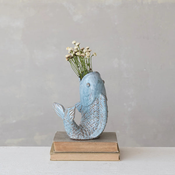 A small blue Fish-Shaped Vase with white flowers, set on two stacked vintage books against a neutral background, brings a touch of coastal home decor.