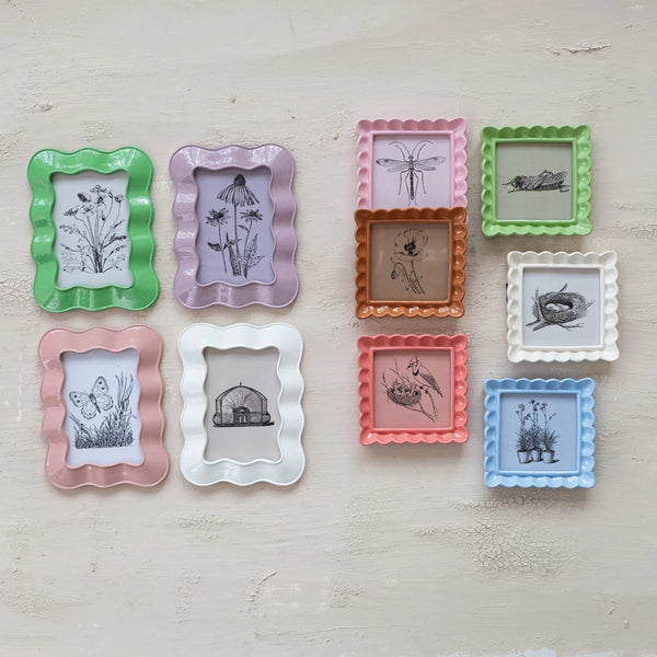 A set of ten Ruffled Enamel Frames in six pastel colors, each displaying a black-and-white botanical or animal sketch, is artfully arranged on a light beige textured wall.